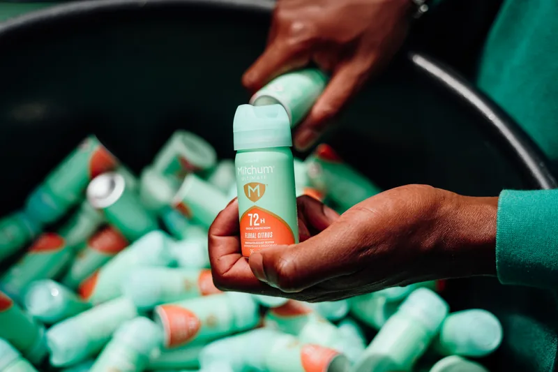 Hands lifting a Mitchum Ultimate antiperspirant from a bin of them at a sampling stand