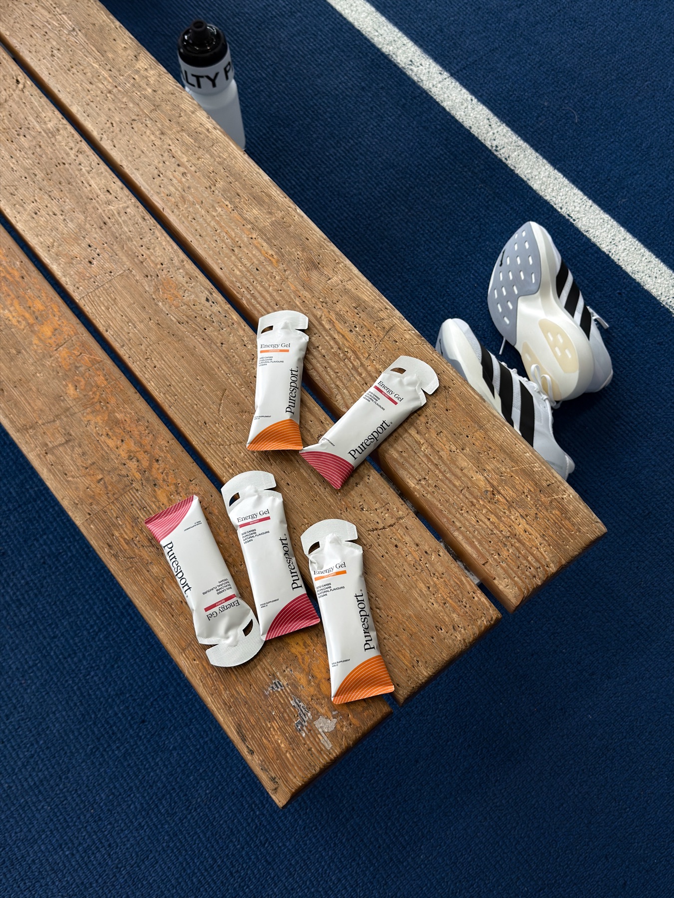 Five Puresport energy gels laid out on a wooden bench beside running spikes at the edge of a blue track